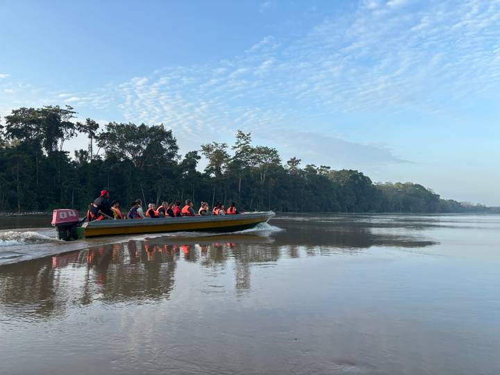 Longboat with life-jacketed tourists speeds along calm rainforest river at dawn.