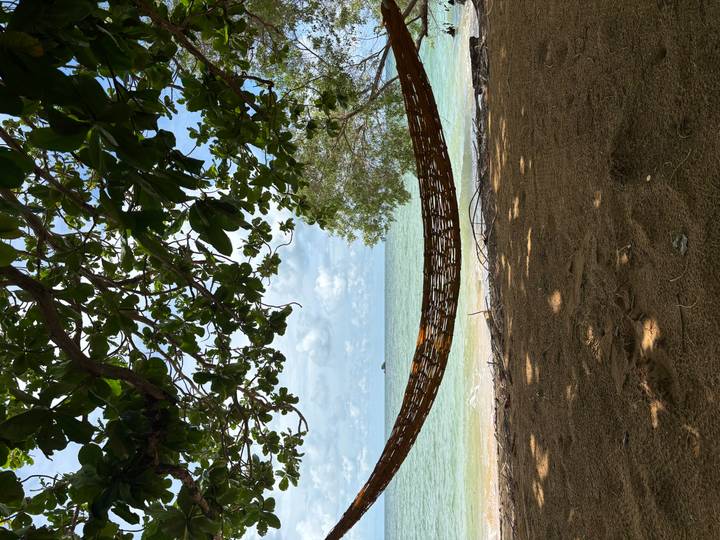 Woven hammock strung under leafy shade facing turquoise sea and distant islet.