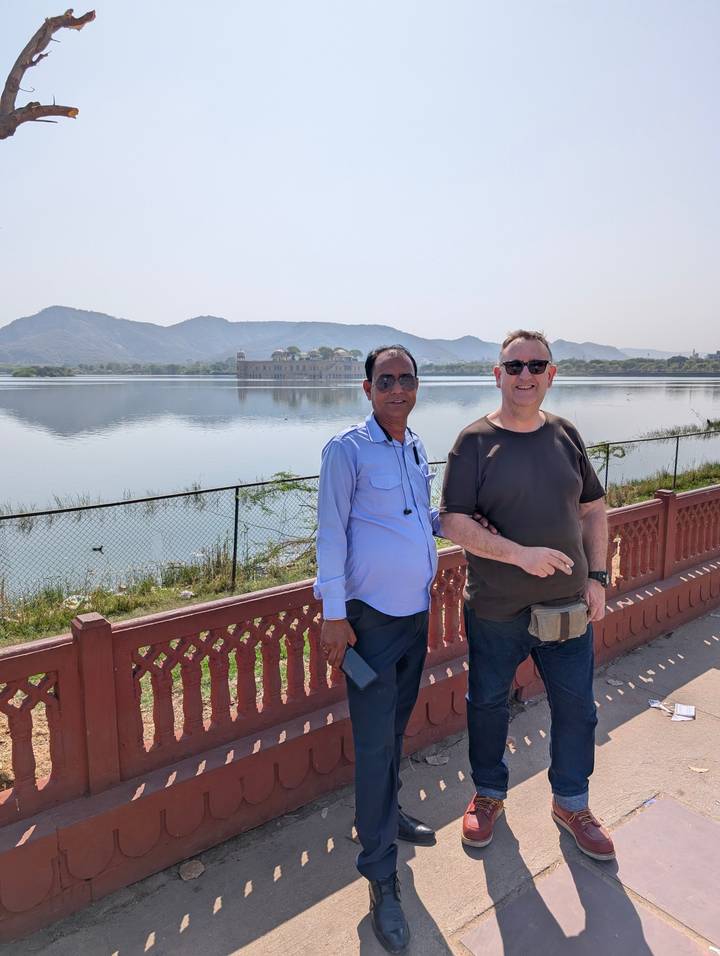 Two men stand beside the calm lake with Jal Mahal palace floating in the distance and arid hills behind.