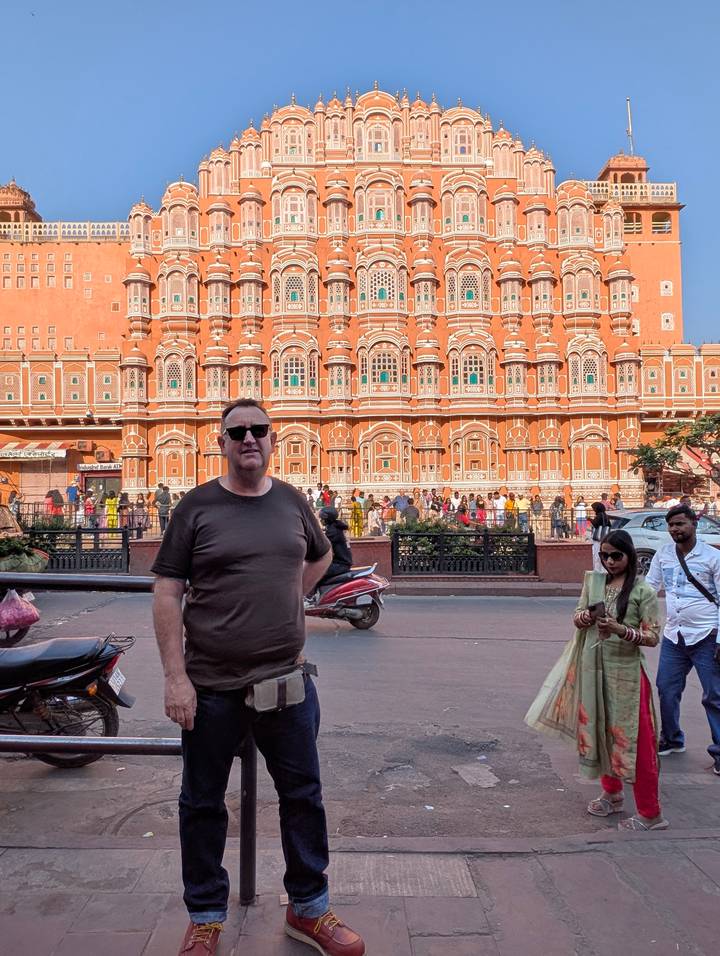 Tourist poses on busy street with the iconic pink facade of Hawa Mahal in Jaipur behind.