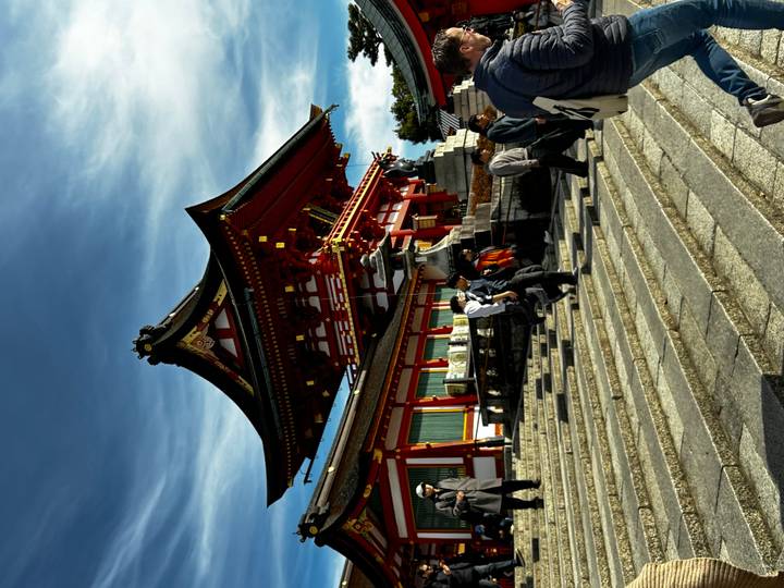 Visitors climb wide stone steps toward a vivid red and white shrine pavilion under dramatic sky.