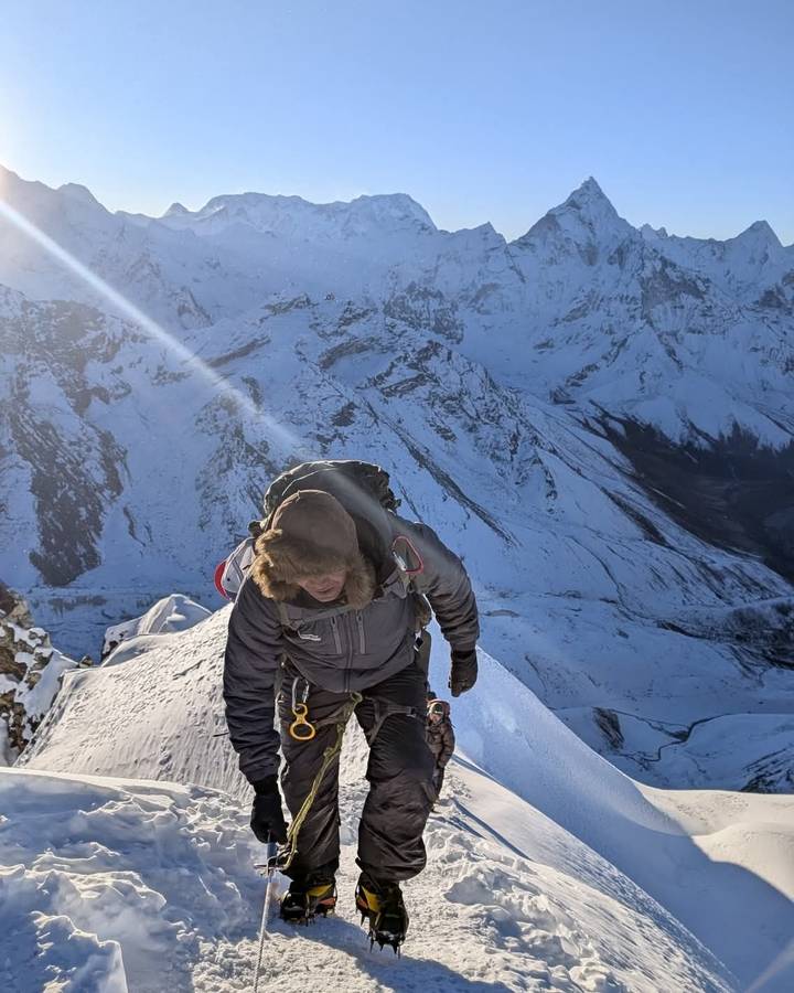 Mountaineer in heavy winter gear climbing a narrow icy ridge with dramatic snow-covered peaks behind.