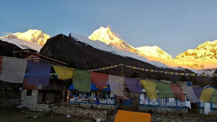 Colorful prayer flags flutter outside a small Himalayan village with sunrise light on snowy peaks.
