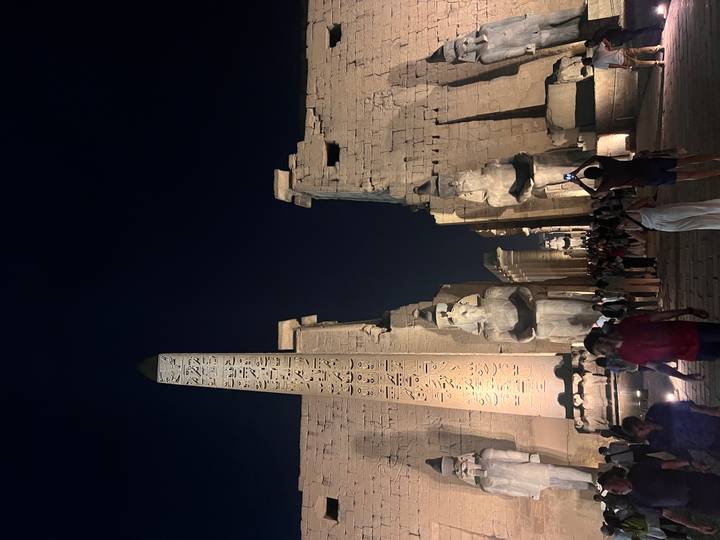 Crowded night scene at Luxor Temple featuring the illuminated obelisk and seated pharaoh statues flanking the grand entrance.