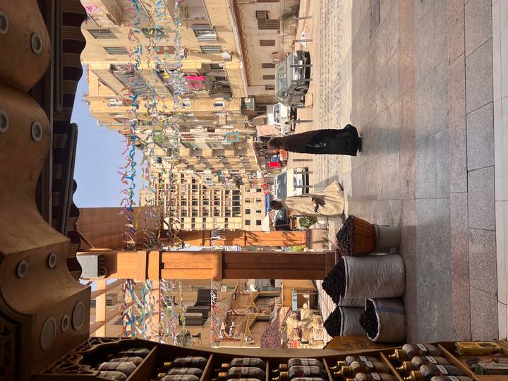 Daytime street scene in an Egyptian city with two locals in traditional clothing walking past market stalls and apartment blocks decorated with colorful streamers.