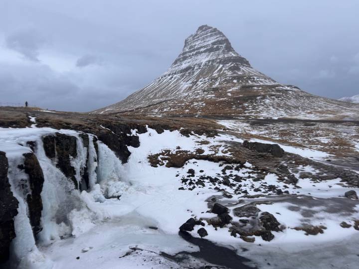 The iconic cone-shaped Kirkjufell mountain rises above a partially frozen waterfall and snow-covered terrain in winter.