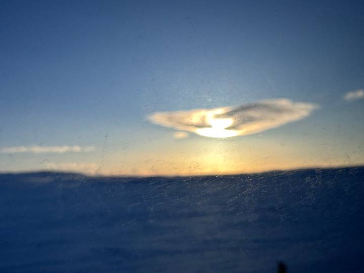 A hazy view of the sun setting behind distant snow-covered land, seen through a scratched window.