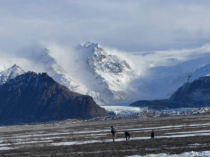 Jagged snow-capped peaks and sweeping glacier tongues rise above a black sand plain where tiny hikers traverse the landscape.