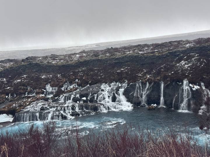 Blue-tinged water cascades in countless icy rivulets over dark basalt ledges at Hraunfossar waterfall on a grey winter day.