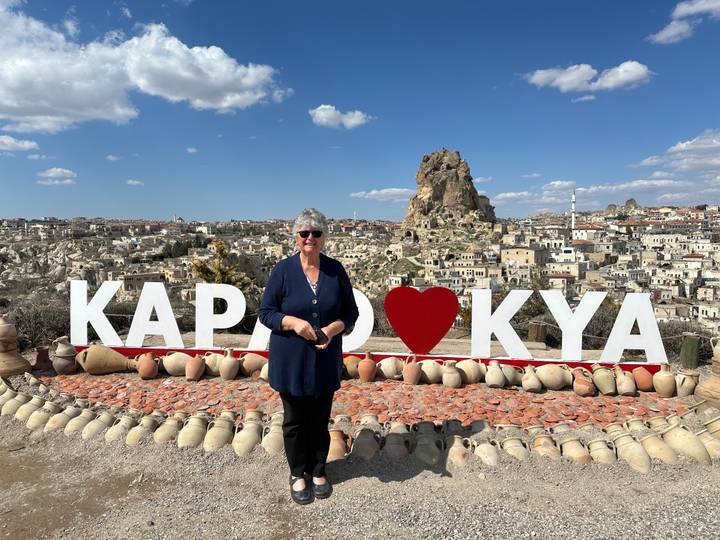 Woman poses before large KAPADOKYA sign with Cappadocia rock citadel rising behind.
