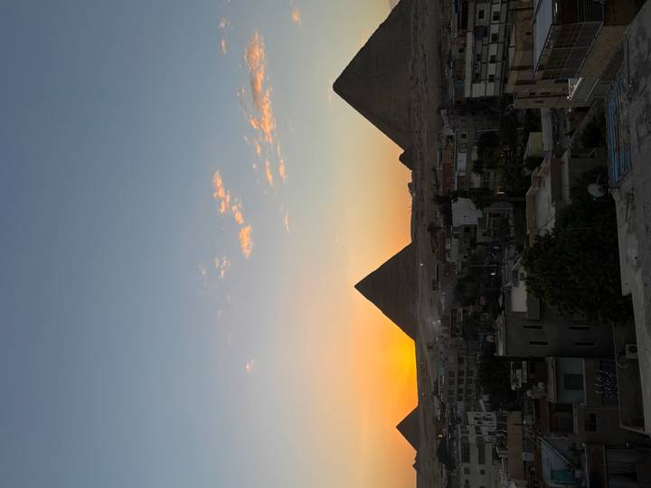 Silhouetted pyramids of Giza against an orange sunset sky above the outskirts of Cairo.