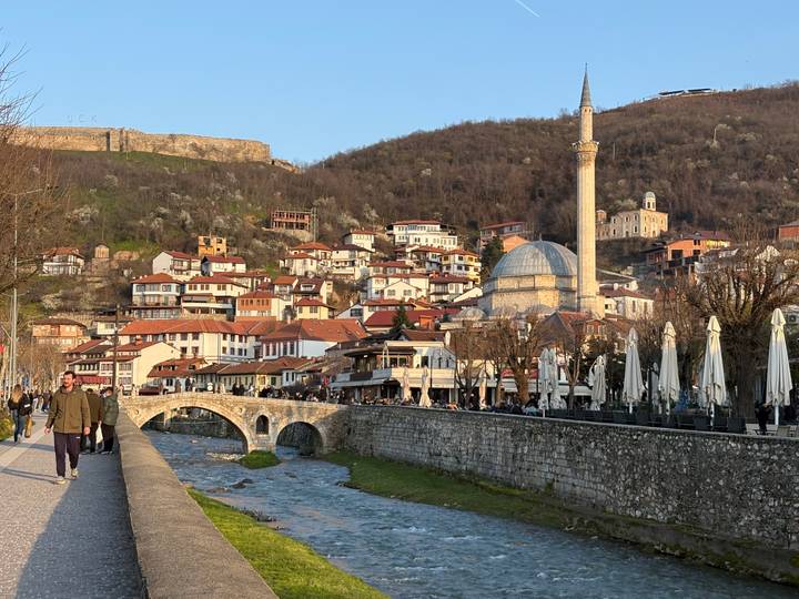 Scenic riverside view of Prizren with Ottoman bridge, mosque, and hillside houses at golden hour.