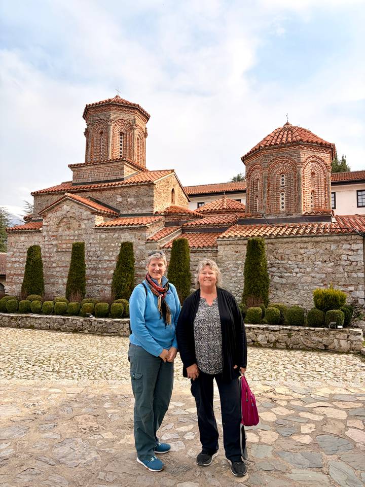 Two women posing happily in front of a stone-built Byzantine monastery with red tiled roofs.