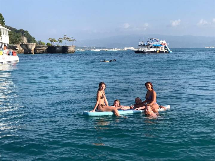 Group of friends on a paddle board and in the water enjoying a turquoise sea with tour boats in the distance.