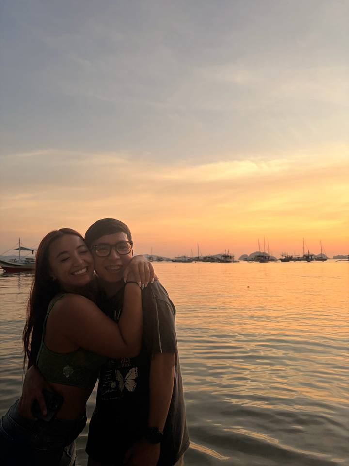 Smiling young couple embracing by the water with yachts silhouetted against a colorful sunset sky.