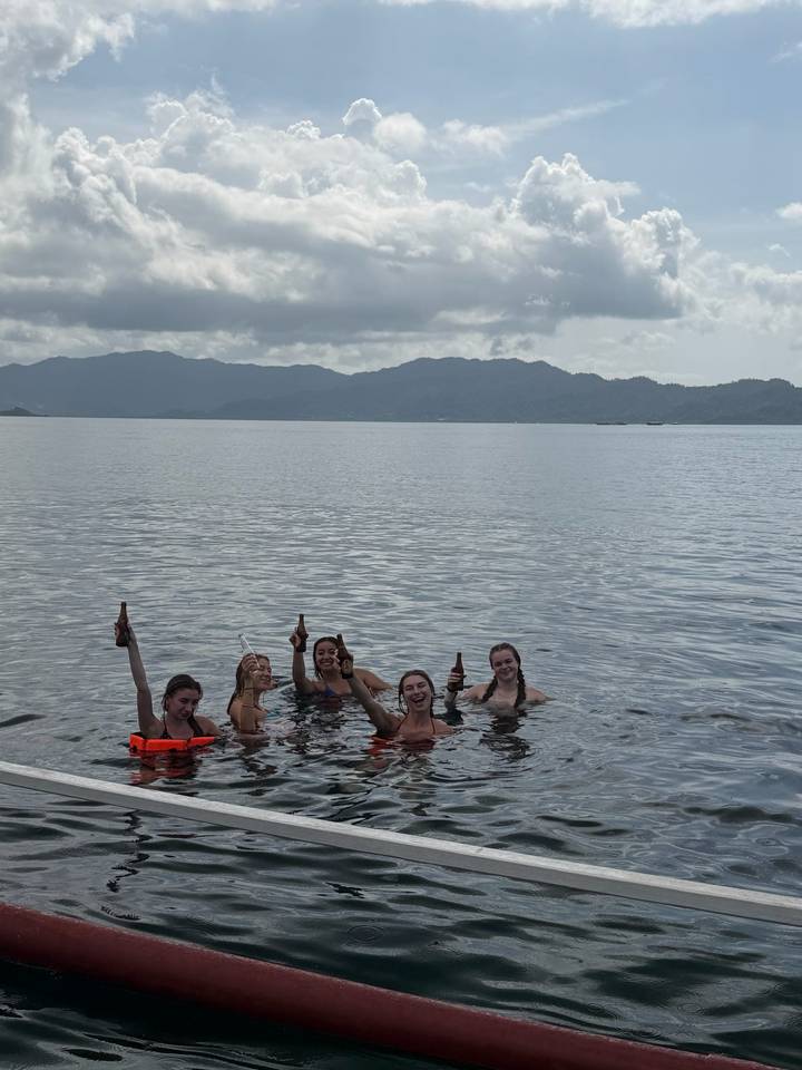 Five friends waist-deep in the sea raising beer bottles with hazy mountains in the background.