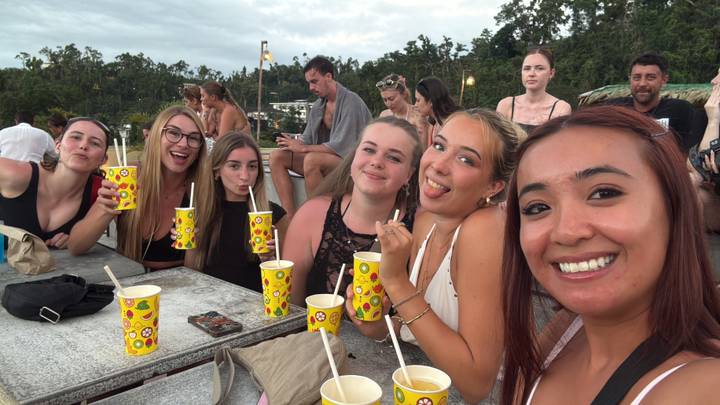 Selfie of smiling women sipping drinks from bright yellow cups at an outdoor wooden table.