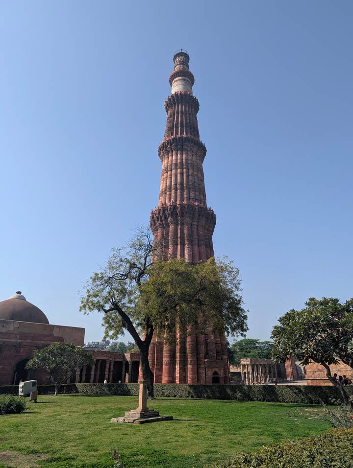 Towering Qutub Minar rising above surrounding trees against a clear blue Delhi sky.