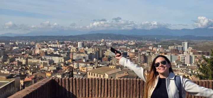 Smiling woman with arms raised overlooking a sprawling Mediterranean cityscape from a terrace.