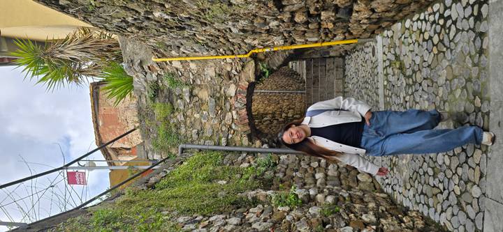Woman walking through a cobblestone alley framed by stone arches and moss-covered walls.