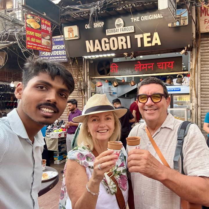 Three travellers holding clay cups of chai and smiling in front of a busy Indian street stall.