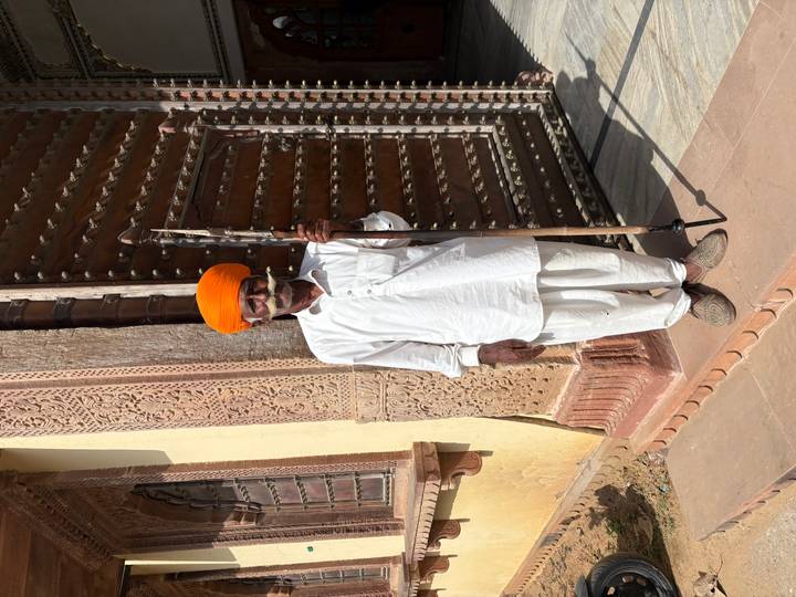 Portrait of a Rajasthani guard in white traditional attire and bright orange turban standing by a carved wooden doorway.