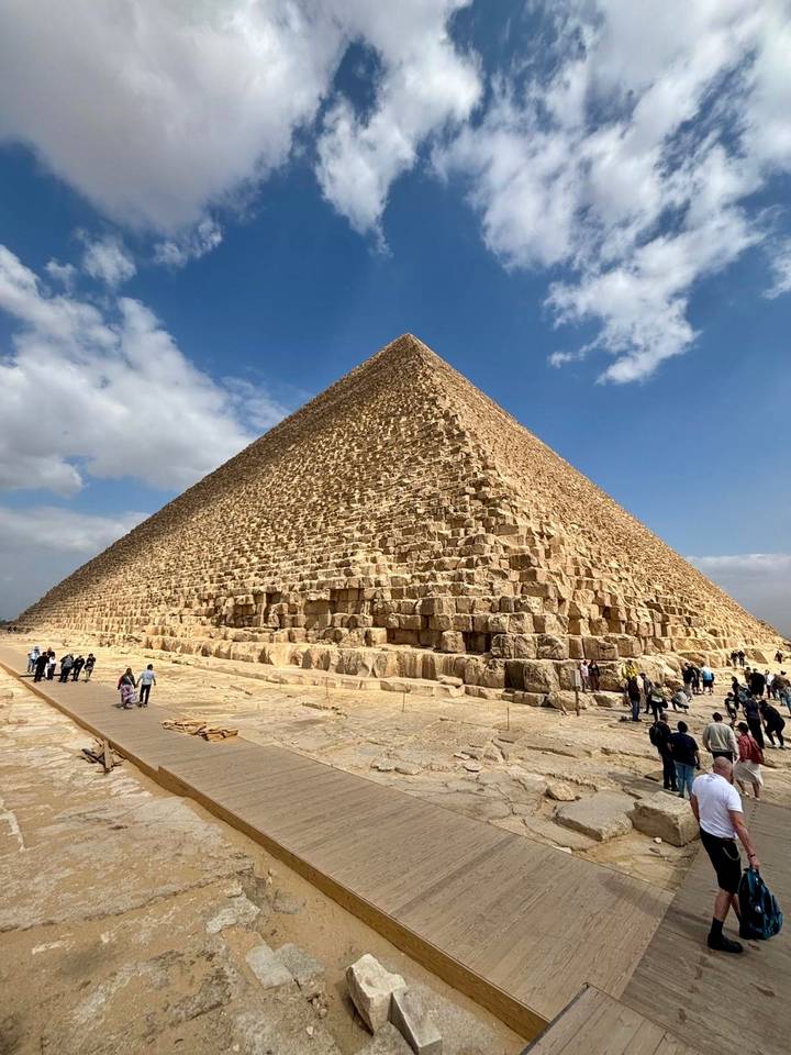Dramatic low-angle view of the Great Pyramid of Giza with tiny visitors at its base against a blue sky with clouds