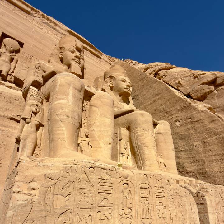 Close-up of the colossal seated statues carved into the sandstone cliff at Abu Simbel under clear blue sky