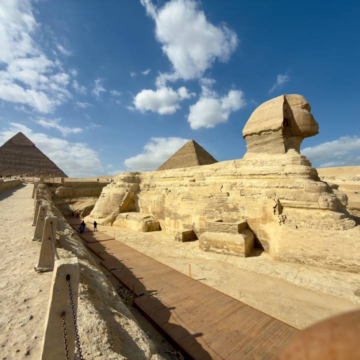 Side profile of the Sphinx with two pyramids rising behind under a bright sky with scattered clouds