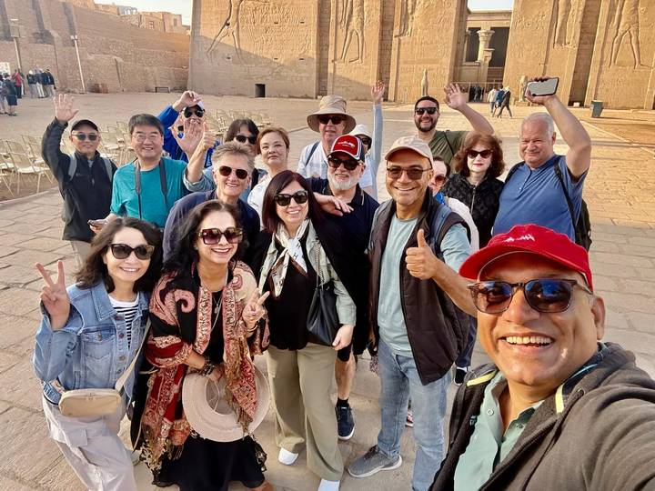 Large selfie group smiling and giving thumbs-up in the courtyard of an Egyptian temple complex.