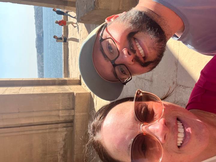 Close selfie of two travellers by ancient stone columns overlooking a wide blue river.