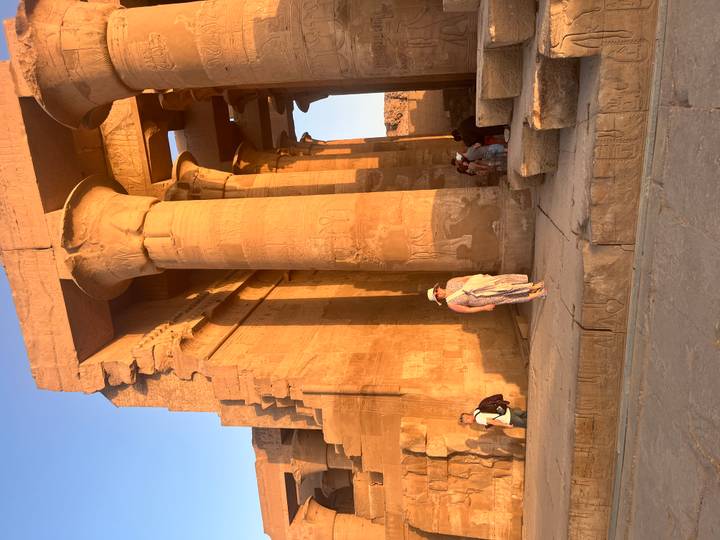 Golden sunlight illuminates towering sandstone columns of an Egyptian temple as visitors stroll between them.