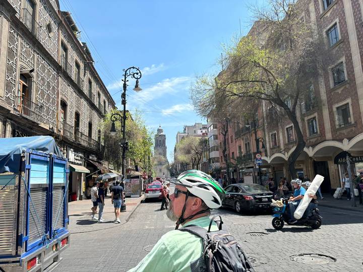 Busy historic street with colonial buildings, pedestrians and a cyclist wearing a helmet in bright sunshine.