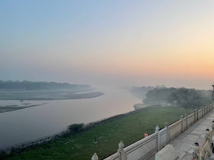 Misty sunrise over a calm river and tree-lined banks creating a tranquil pastel scene.
