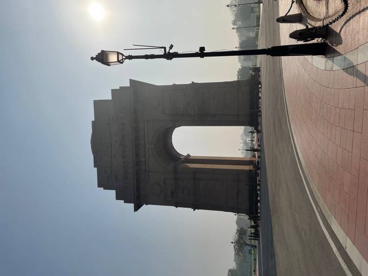 Silhouetted India Gate rises against a hazy blue morning sky on an empty boulevard in Delhi.