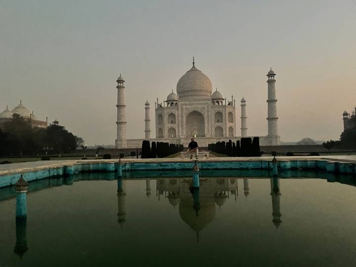 Iconic Taj Mahal reflected in a still pool at dawn with a lone visitor seated on the central platform.