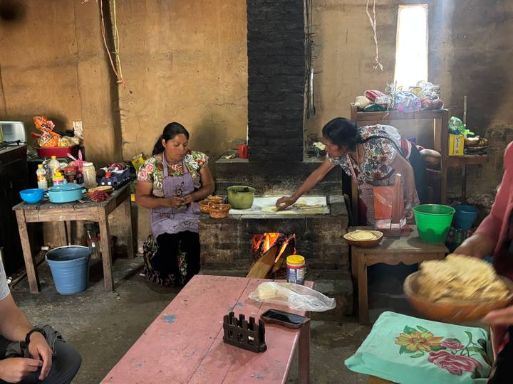 Two indigenous women prepare tortillas over a wood-fired hearth inside a rustic kitchen.