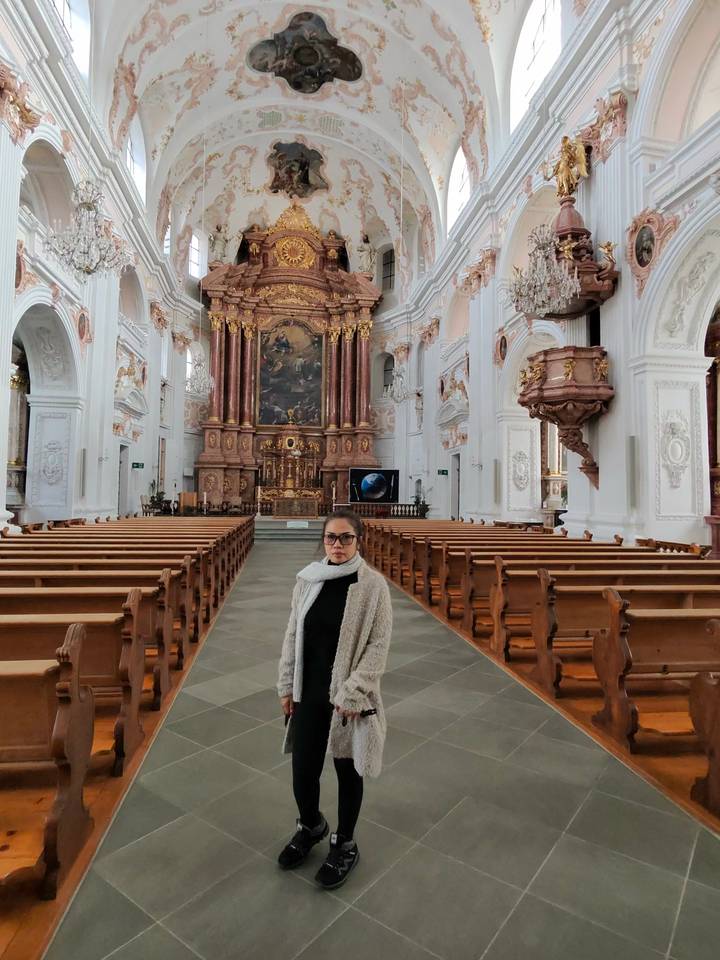 Woman posing inside an ornate baroque church with wooden pews and elaborate altar.
