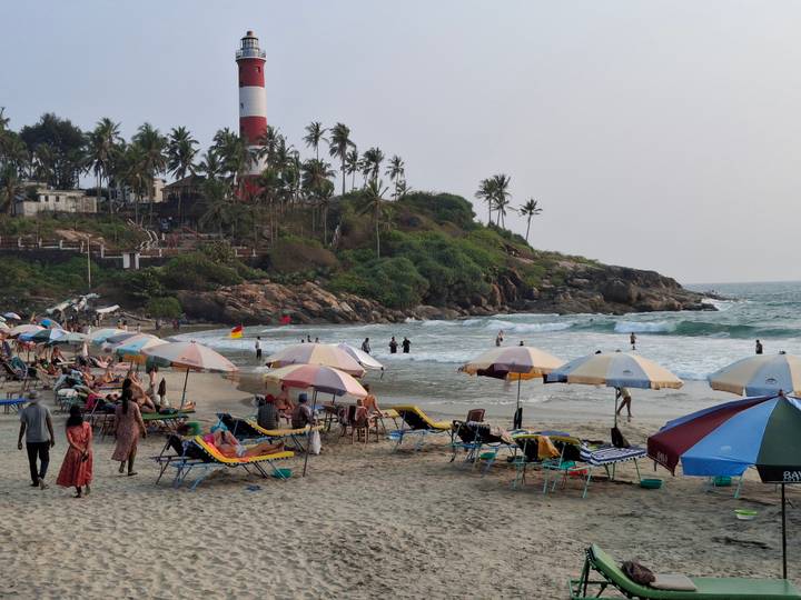 Busy Kovalam beach scene with colorful umbrellas, sunbeds and lighthouse on a rocky headland.