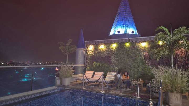 Rooftop swimming pool terrace at night with city lights and a cone-shaped illuminated tower.