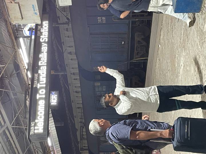 Guide gestures toward illuminated sign reading 'Welcome to Tundla Railway Station' as traveler looks on.