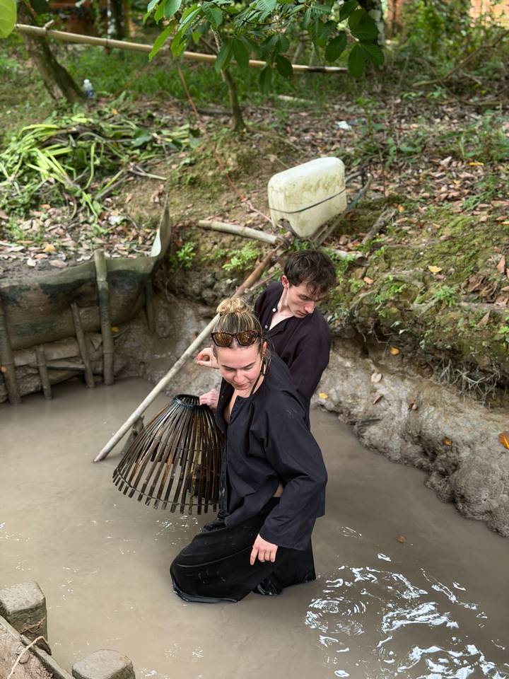 Two travelers wearing dark clothing search for fish with a bamboo basket trap in a muddy canal.