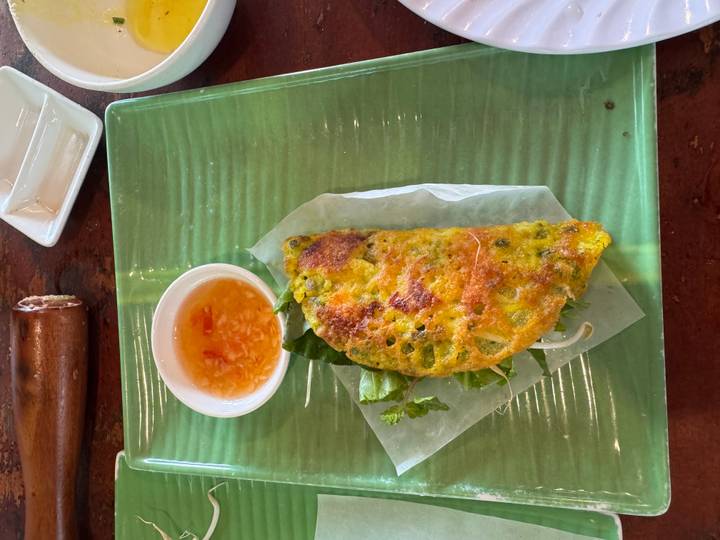 A crispy yellow Vietnamese pancake rests on lettuce with dipping sauce on a green plate.