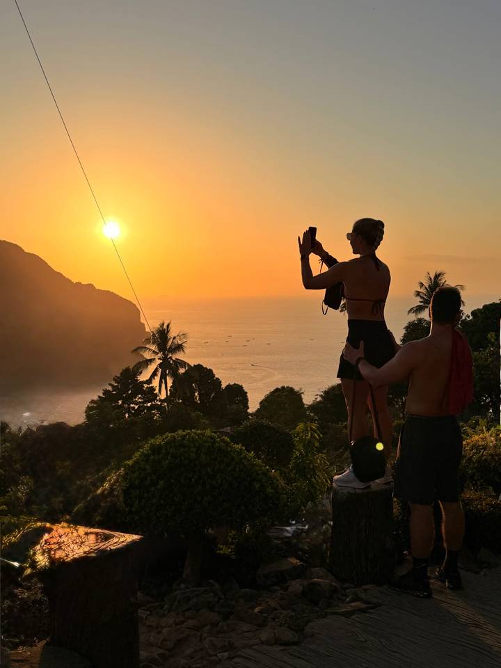 Man supporting woman as she photographs a glowing tropical sunset from a hill viewpoint