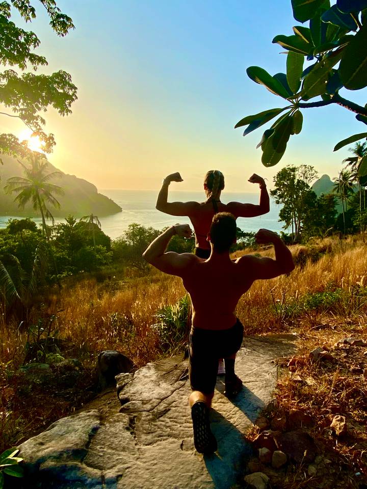 Silhouetted couple flexing their arms on a hilltop viewpoint overlooking a tropical bay at sunset.