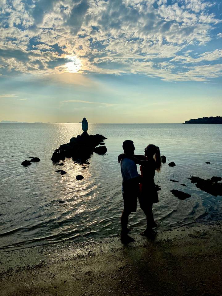 Silhouette of a couple embracing on a rocky shore of a calm sea with a vertical stone sculpture in the distance.