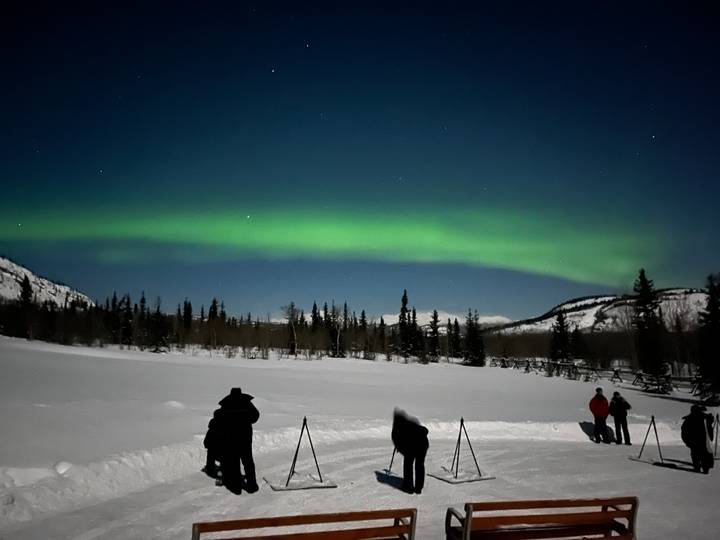 Photographers stand on snow with tripods capturing a sweeping green aurora arc in the night sky