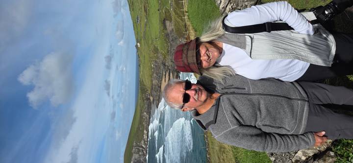 Smiling older couple posing in front of rugged coastal cliffs and crashing waves on a bright day.