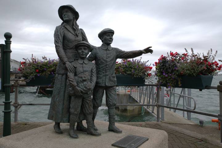 Bronze emigrant family statue on a waterfront promenade decorated with colorful flower boxes on a cloudy day.