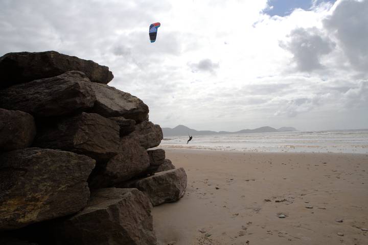 Windy sandy beach with a lone kite-surfer suspended in the air and mountains on the horizon.